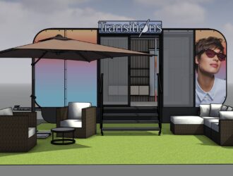 3D rendering of a modern Transitions outdoor exhibition booth featuring a green carpet area with white seating, umbrellas, and large displays showing a person wearing Transitions lenses, set against a cloudy sky backdrop.