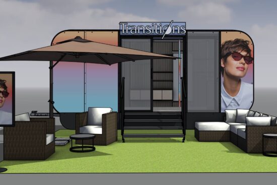 3D rendering of a modern Transitions outdoor exhibition booth featuring a green carpet area with white seating, umbrellas, and large displays showing a person wearing Transitions lenses, set against a cloudy sky backdrop.