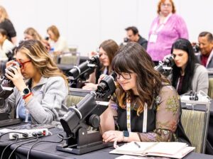 Étudiants utilisant des microscopes dans un laboratoire ou une salle de classe. Au premier plan, une femme avec des lunettes et de longs cheveux bruns portant un haut à motifs floraux regarde dans un microscope tout en prenant des notes. À sa gauche, une autre étudiante aux cheveux blonds utilise également un microscope. Plusieurs autres étudiants sont visibles en arrière-plan travaillant à des paillasses de laboratoire avec des microscopes et de l'équipement. Une instructrice en chemise rose se tient en arrière-plan supervisant la séance.