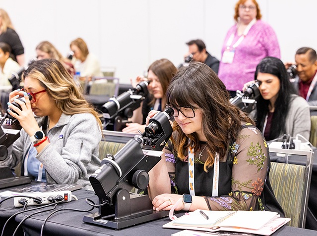 Étudiants utilisant des microscopes dans un laboratoire ou une salle de classe. Au premier plan, une femme avec des lunettes et de longs cheveux bruns portant un haut à motifs floraux regarde dans un microscope tout en prenant des notes. À sa gauche, une autre étudiante aux cheveux blonds utilise également un microscope. Plusieurs autres étudiants sont visibles en arrière-plan travaillant à des paillasses de laboratoire avec des microscopes et de l'équipement. Une instructrice en chemise rose se tient en arrière-plan supervisant la séance.