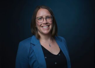 Headshot of Dr. Allison Scott, a woman with shoulder-length brown hair and glasses, smiling warmly at the camera. She's wearing a blue blazer over a black top with small decorative details and a pendant necklace, photographed against a dark teal background.