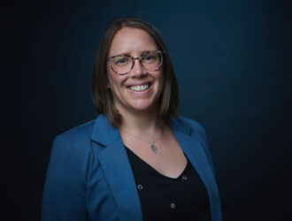 Headshot of Dr. Allison Scott, a woman with shoulder-length brown hair and glasses, smiling warmly at the camera. She's wearing a blue blazer over a black top with small decorative details and a pendant necklace, photographed against a dark teal background.