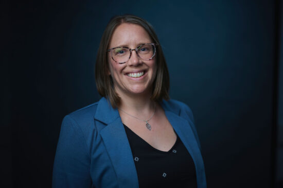 Headshot of Dr. Allison Scott, a woman with shoulder-length brown hair and glasses, smiling warmly at the camera. She's wearing a blue blazer over a black top with small decorative details and a pendant necklace, photographed against a dark teal background.