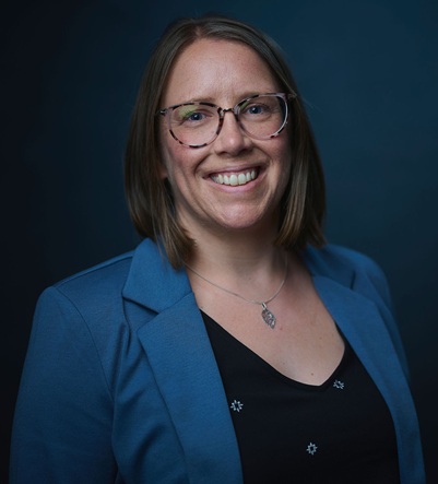 Portrait du Dr. Allison Scott, une femme aux cheveux bruns mi-longs et portant des lunettes, souriant chaleureusement à l'appareil photo. Elle porte un blazer bleu sur un haut noir avec de petits détails décoratifs et un collier avec pendentif, photographiée sur un fond bleu-vert foncé.
