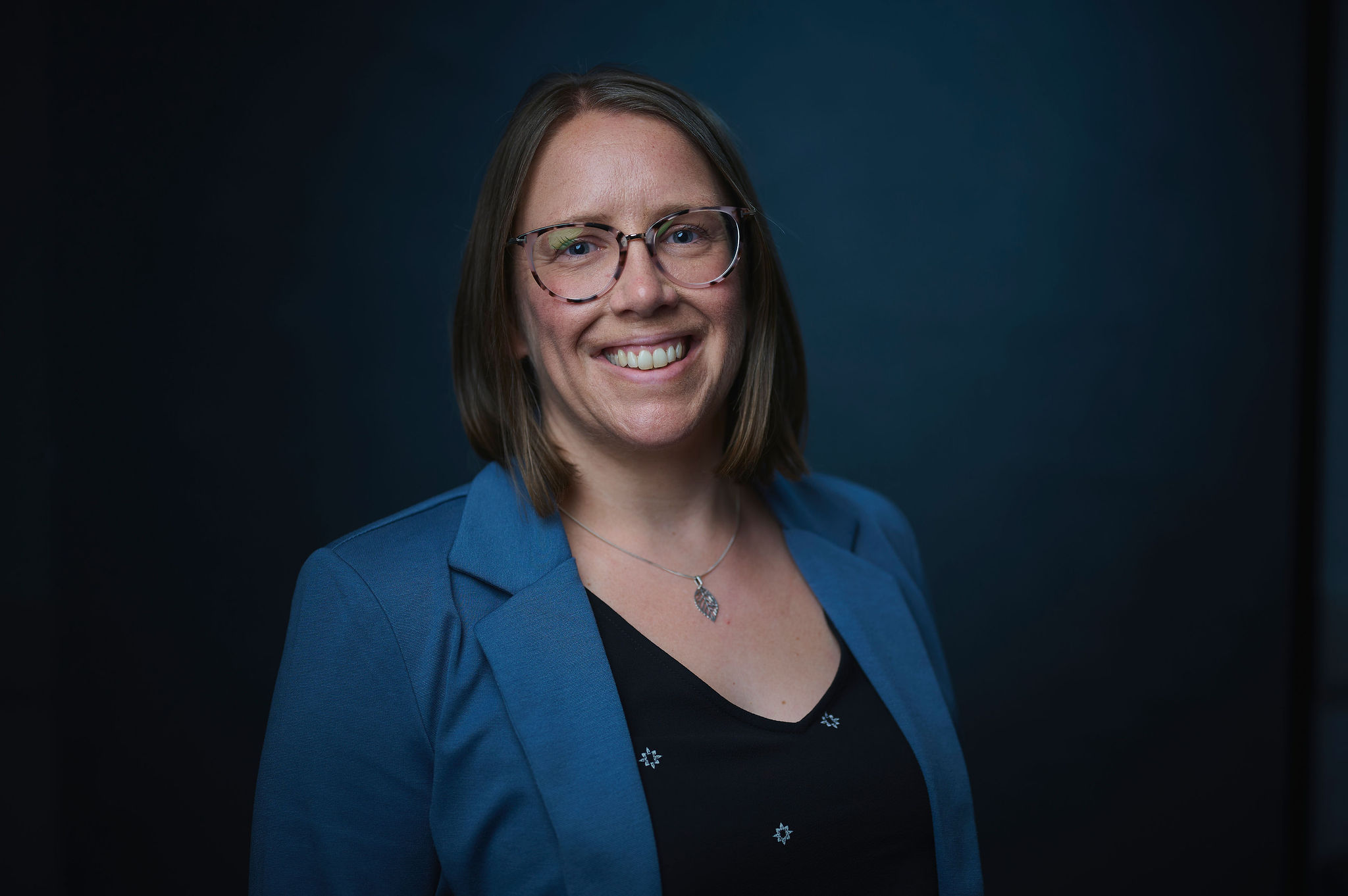 Headshot of Dr. Allison Scott, a woman with shoulder-length brown hair and glasses, smiling warmly at the camera. She's wearing a blue blazer over a black top with small decorative details and a pendant necklace, photographed against a dark teal background.