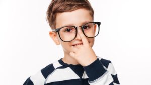 A young boy with brown hair wearing black-rimmed glasses and a navy and white striped sweater, thoughtfully placing his finger near his chin while smiling at the camera.