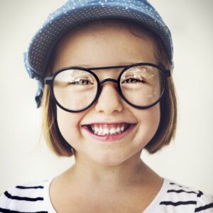 A young girl with short brown hair wearing oversized black round glasses and a blue polka-dot hat, smiling widely at the camera. She's wearing a white shirt with black stripes.