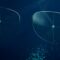 Main article image: Image of two translucent eyeglass lenses floating in a dark blue digital space, surrounded by scattered particles of light and geometric grid patterns. The image suggests a smart lens technology with a high-tech, futuristic aesthetic