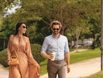 Un couple marchant ensemble à l'extérieur par une journée ensoleillée, tous deux portant des lunettes de soleil. La femme porte une robe fluide de couleur pêche avec un petit sac à main, et l'homme porte une chemise bleu clair avec un pantalon marron tout en tenant une tasse de café. Ils se promènent dans un parc bordé d'arbres avec de l'herbe verte et du feuillage en arrière-plan.