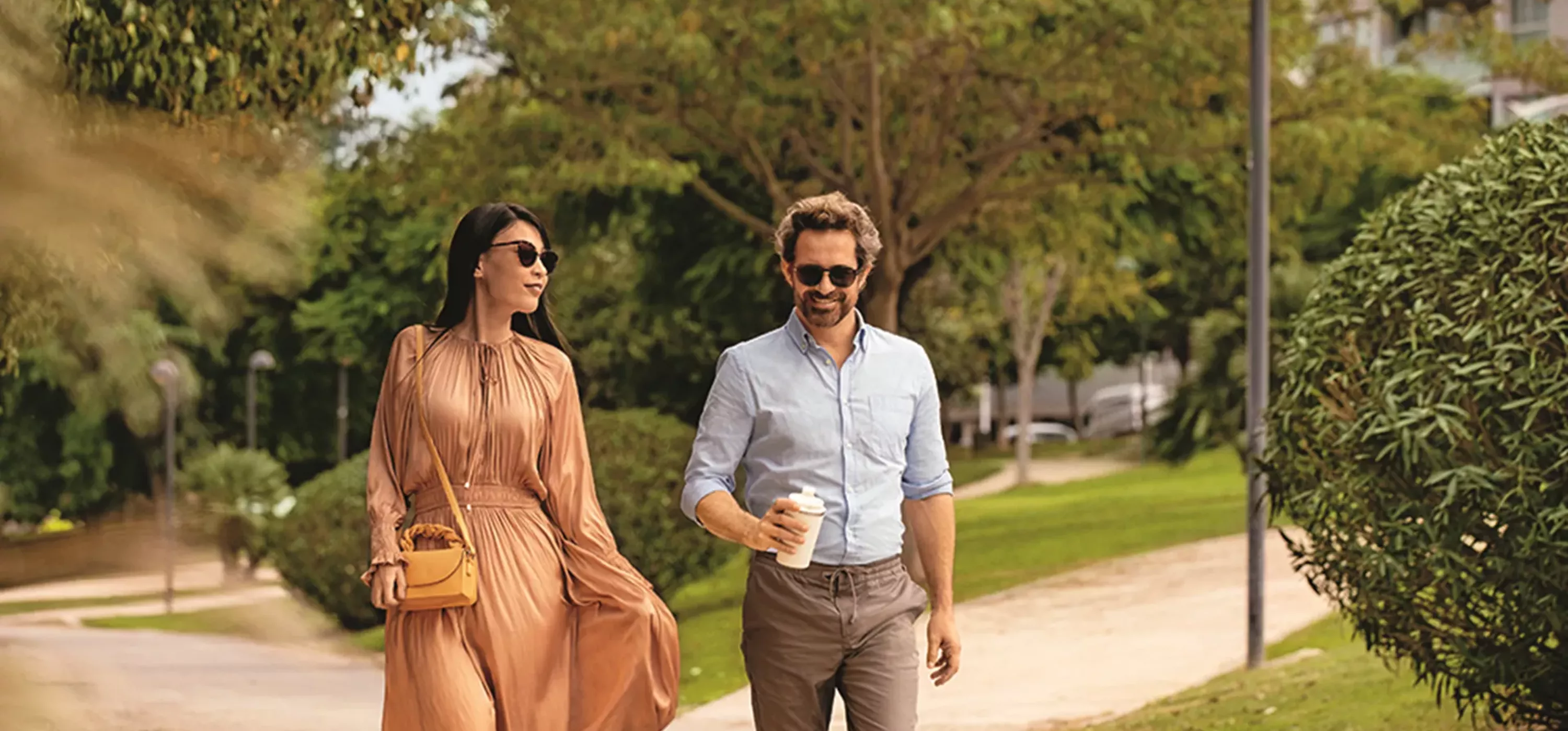 A couple walking together outdoors on a sunny day, both wearing sunglasses. The woman is dressed in a flowing peach-colored dress with a small handbag, and the man is wearing a light blue shirt with brown pants while holding a coffee cup. They are strolling through a tree-lined park area with green grass and foliage in the background.