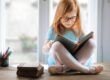 A young girl with long blonde hair and glasses sitting cross-legged on a wooden table, reading a book. She's wearing a light blue t-shirt, with colored pencils and other books visible nearby by a bright window.
