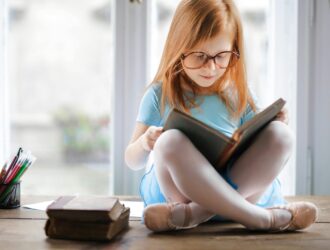 A young girl with long blonde hair and glasses sitting cross-legged on a wooden table, reading a book. She's wearing a light blue t-shirt, with colored pencils and other books visible nearby by a bright window.