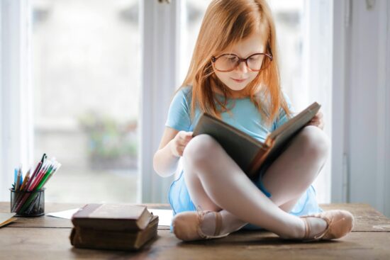 A young girl with long blonde hair and glasses sitting cross-legged on a wooden table, reading a book. She's wearing a light blue t-shirt, with colored pencils and other books visible nearby by a bright window.
