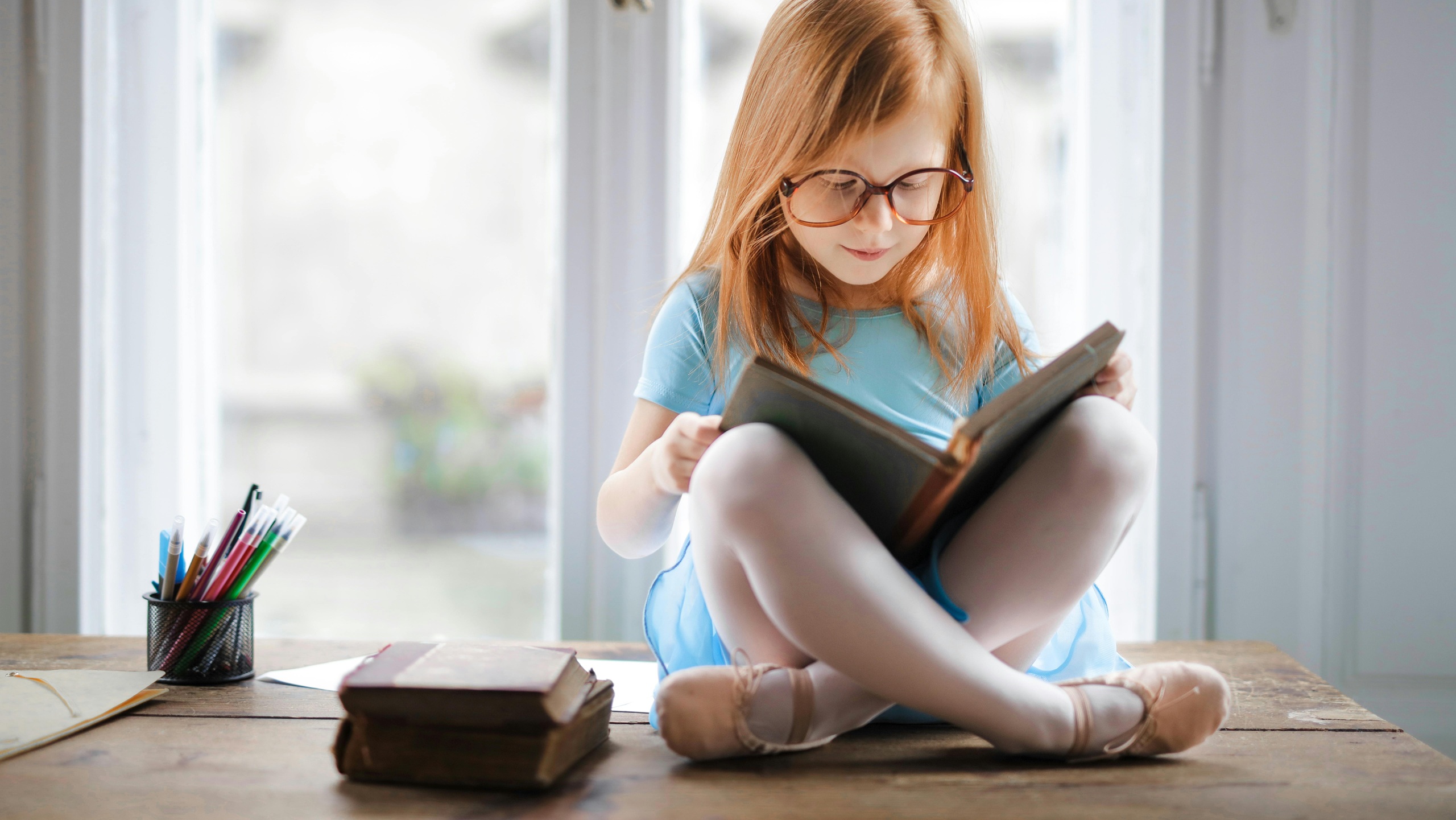 A young girl with long blonde hair and glasses sitting cross-legged on a wooden table, reading a book. She's wearing a light blue t-shirt, with colored pencils and other books visible nearby by a bright window.