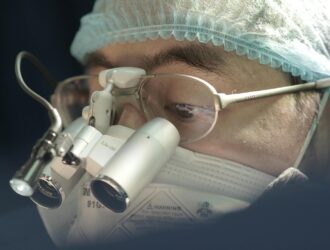 Ophthalmologist performing eye surgery using a surgical microscope, wearing protective surgical cap and glasses in a sterile operating room environment