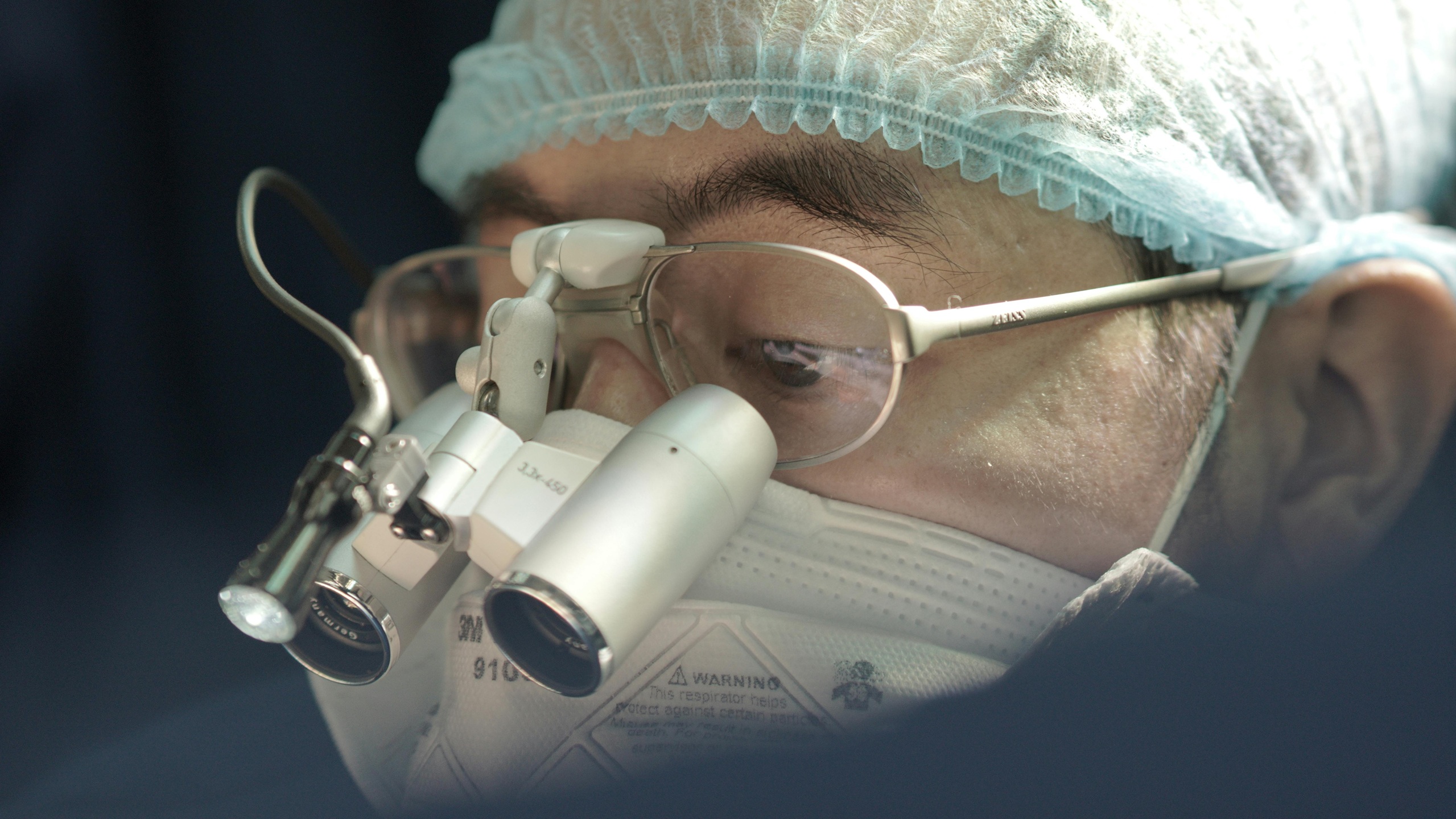 Ophthalmologist performing eye surgery using a surgical microscope, wearing protective surgical cap and glasses in a sterile operating room environment