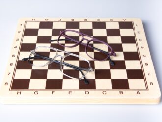 Deux paires de lunettes de la collection LOOK disposées artistiquement sur un échiquier en bois - une paire en gris-bleu translucide et une autre en violet profond, démontrant la précision stratégique et la philosophie de design réfléchie de l'artisanat italien de lunetterie.
