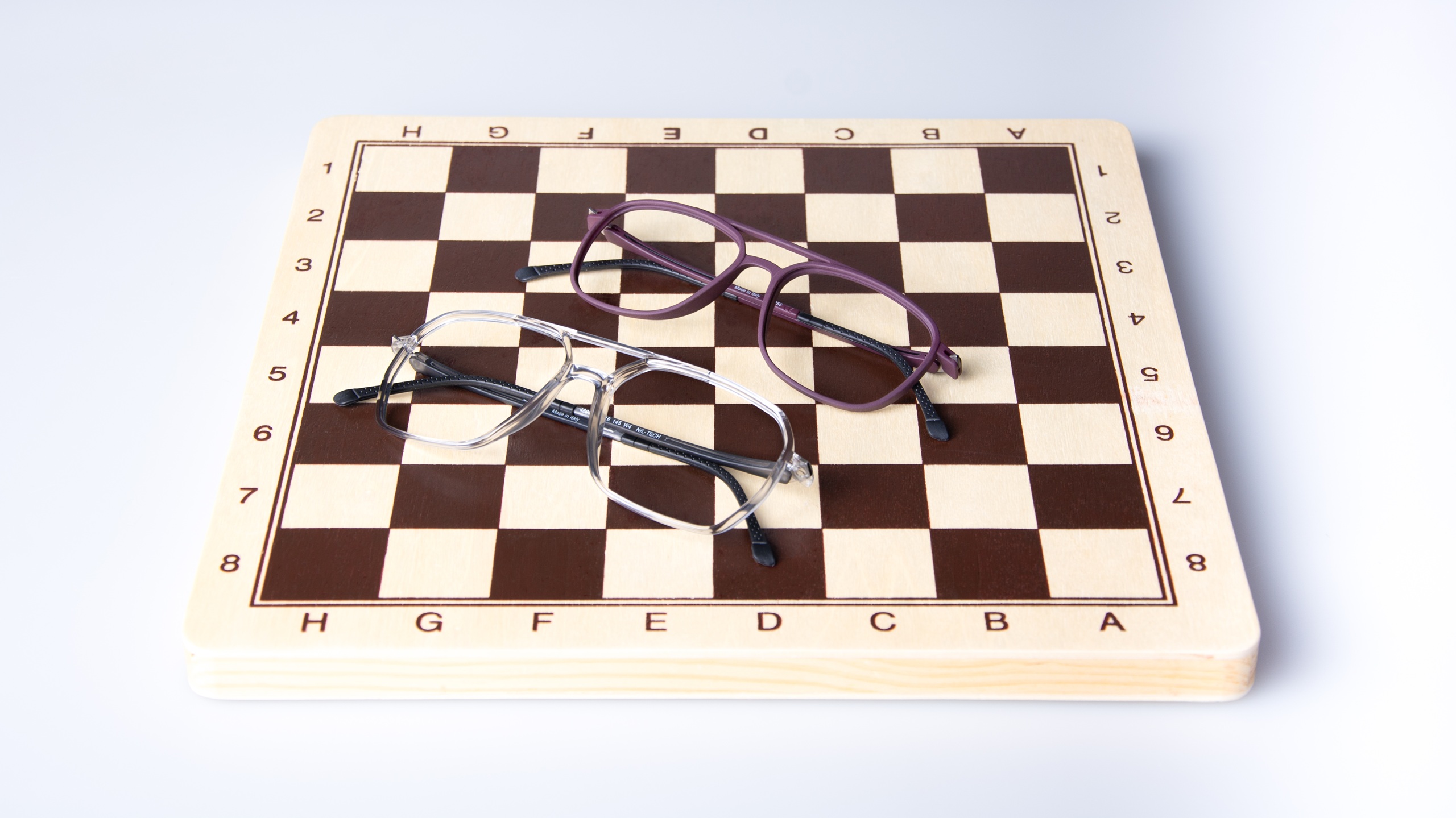 Two pairs of eyeglasses from the LOOK collection placed artistically on a wooden chess board - one pair in translucent gray-blue and another in deep purple, demonstrating the strategic precision and thoughtful design philosophy of Italian eyewear craftsmanship.