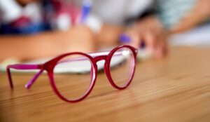 A pair of red-framed eyeglasses sits on a wooden desk surface with a blurred background showing hands and eyecare equipment, representing DOT™ lenses for children's myopia control.