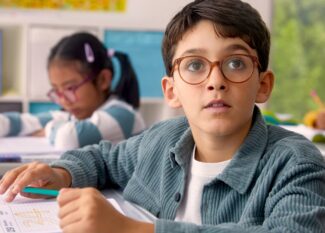 A young boy wearing brown-framed glasses looks up thoughtfully while working at his desk in a classroom setting, with other students visible in the background, showcasing a child with DOT™ lenses engaged in learning.
