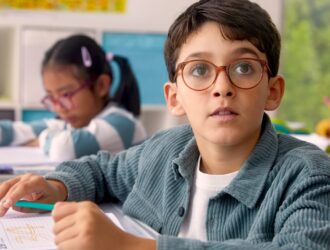 A young boy wearing brown-framed glasses looks up thoughtfully while working at his desk in a classroom setting, with other students visible in the background, showcasing a child with DOT™ lenses engaged in learning.