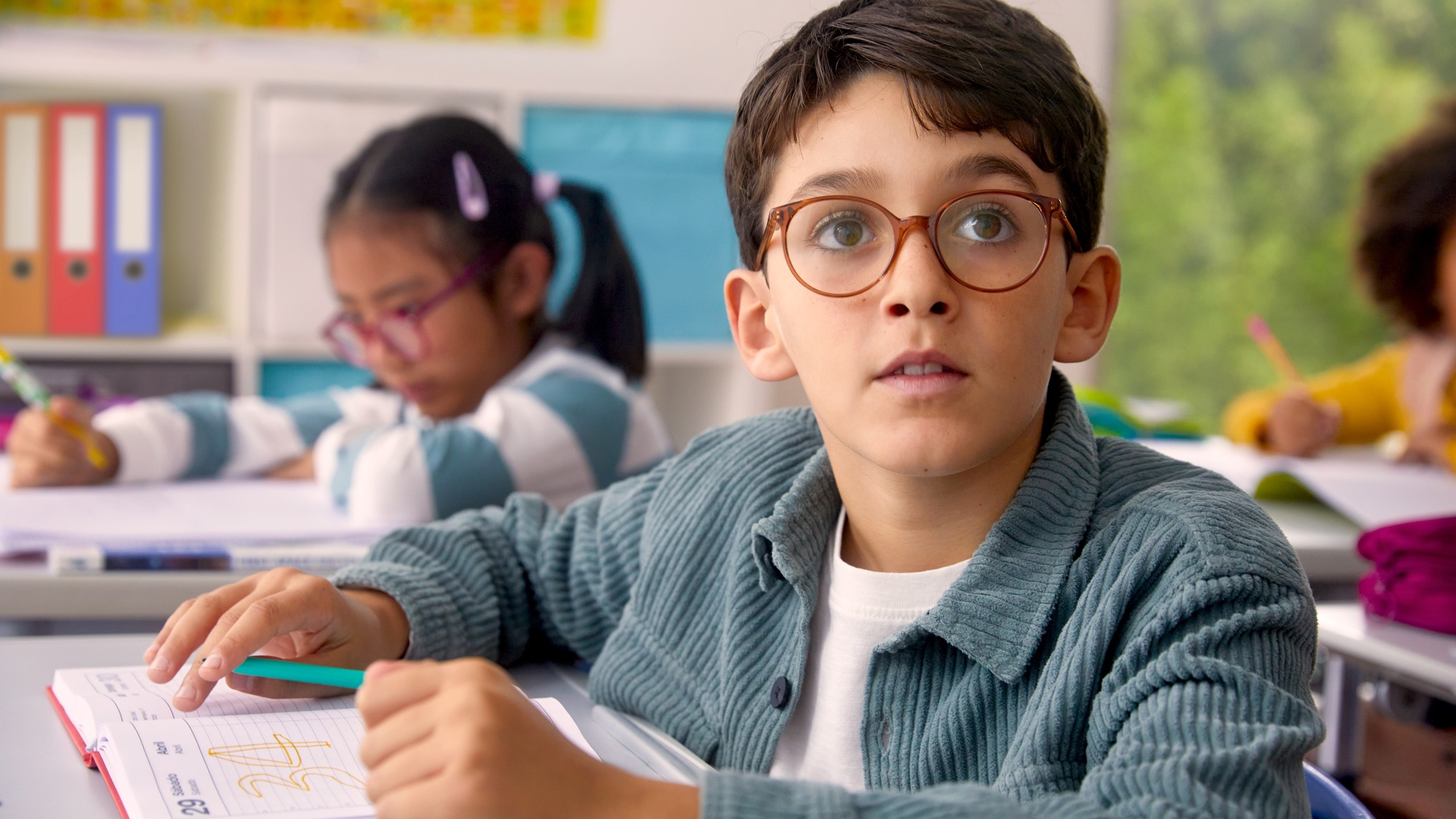 Un jeune garçon portant des lunettes à monture brune lève les yeux pensivement en travaillant à son bureau dans une salle de classe, avec d'autres élèves visibles en arrière-plan, montrant un enfant avec des verres DOT™ engagé dans l'apprentissage.