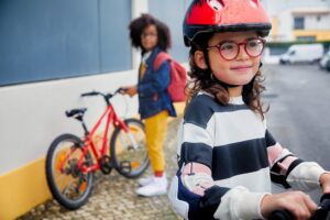 A young girl wearing red-framed glasses and a red bicycle helmet smiles at the camera while sitting on a scooter, with another child on a bicycle visible in the background, demonstrating active children using DOT™ lenses for myopia control.