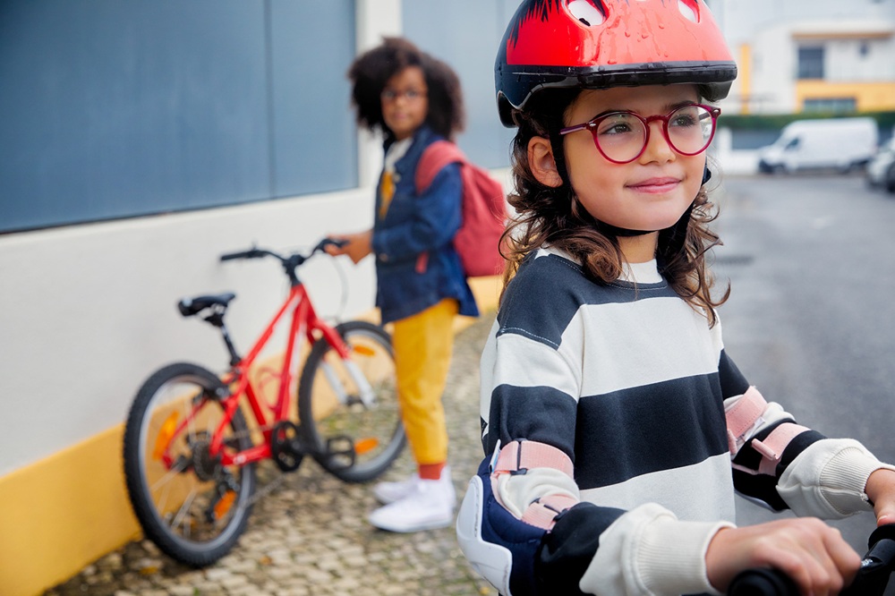 Une jeune fille portant des lunettes à monture rouge et un casque de vélo rouge sourit à la caméra en étant assise sur une trottinette, avec un autre enfant sur un vélo visible en arrière-plan, démontrant des enfants actifs utilisant les verres DOT™ pour le contrôle de la myopie.