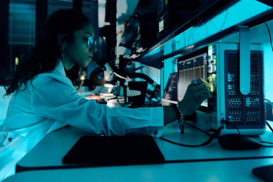 Une chercheuse en blouse blanche travaille à un poste de travail high-tech avec plusieurs moniteurs d'ordinateur et équipements scientifiques dans un environnement de laboratoire moderne, éclairé par une lumière bleue, représentant les activités de recherche et développement du Smart Eyewear Lab d'EssilorLuxottica.