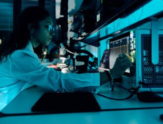 A female researcher in a white lab coat works at a high-tech workstation with multiple computer monitors and scientific equipment in a modern laboratory setting, illuminated by blue lighting, representing EssilorLuxottica's Smart Eyewear Lab research and development activities.
