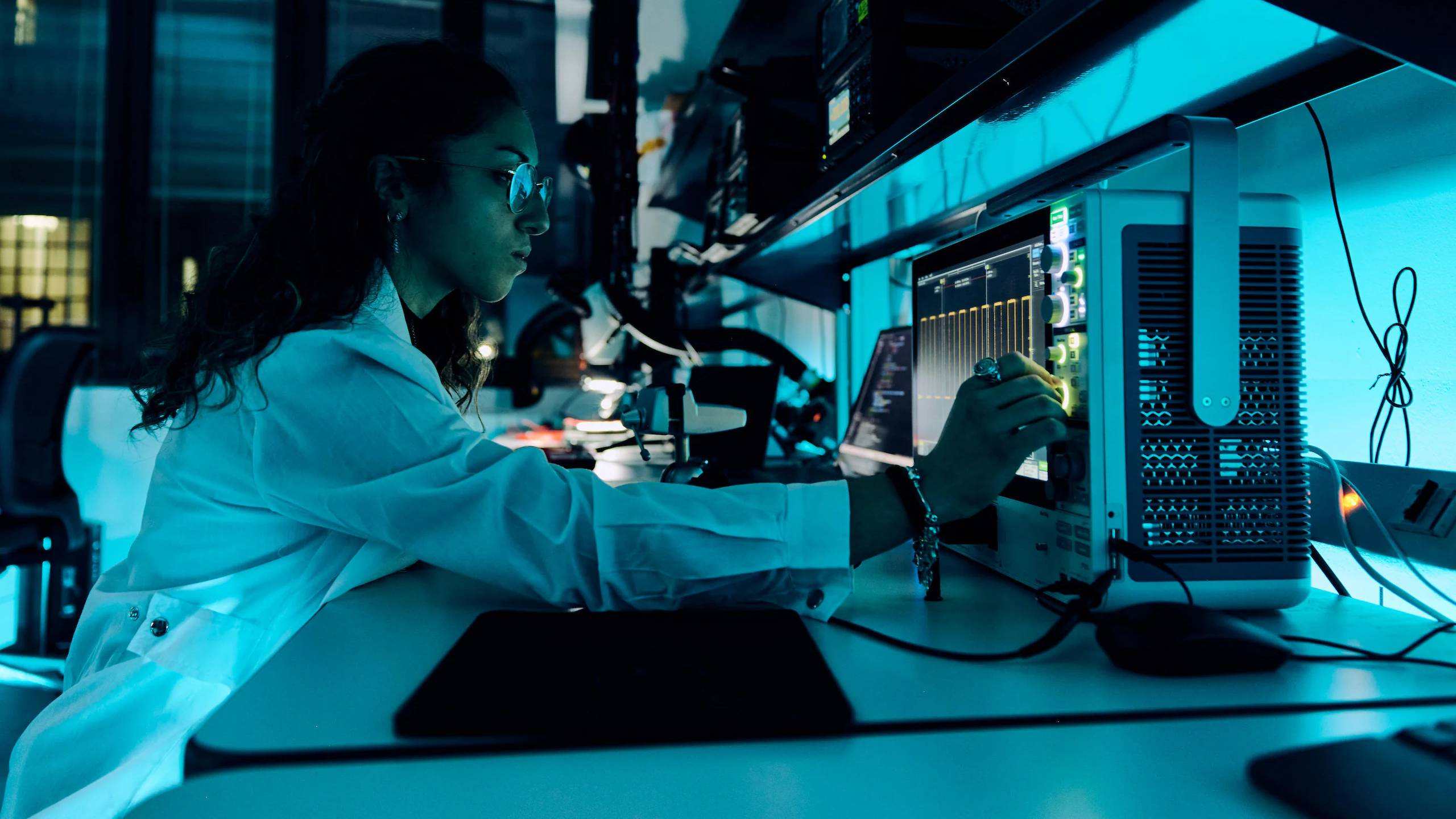 A female researcher in a white lab coat works at a high-tech workstation with multiple computer monitors and scientific equipment in a modern laboratory setting, illuminated by blue lighting, representing EssilorLuxottica's Smart Eyewear Lab research and development activities.