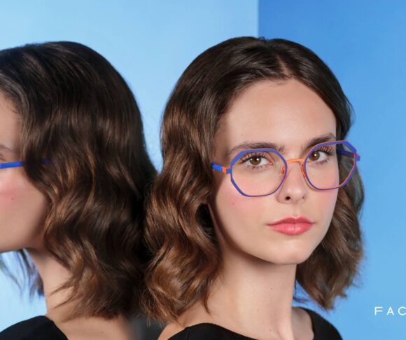Visuel de campagne Face à Face, montrant une femme portant des lunettes géométriques colorées bleu et orange, posant à côté d’un miroir reflétant son profil sur un fond bleu dégradé.