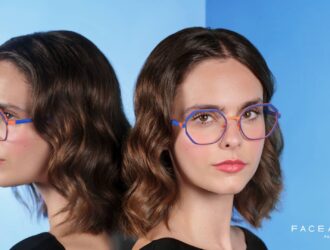 Campaign image for Face à Face, showing a woman wearing colorful geometric eyeglasses in blue and orange, standing beside a mirror that reflects her profile against a gradient blue background.