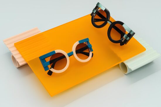 Artistic display of FACE A FACE sunglasses on colorful acrylic platforms - two pairs of avant-garde eyewear with geometric frames in pink, blue, and black colors arranged on bright orange, peach, and mint green translucent surfaces.