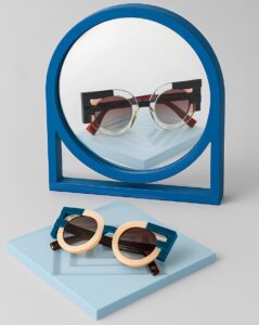 Modern eyewear display featuring FACE A FACE sunglasses with a blue circular mirror stand - geometric sunglasses with peach and blue color-blocking frames displayed on light blue platforms alongside a contemporary blue mirror holder.