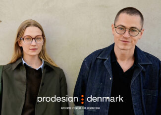 Two people wearing Prodesign Denmark eyewear standing against a concrete wall. On the left, a woman with blonde hair wearing round tortoiseshell glasses and an olive green jacket. On the right, a person with short hair wearing round black-framed glasses and a dark denim jacket. The Prodesign Denmark logo and tagline 'Authentic Eyewear for Generations' appears at the bottom.