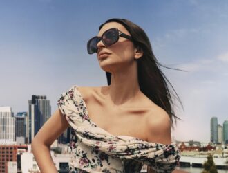 Emily Ratajkowski portant des lunettes de soleil Tory Burch "Pierced" noires avec des détails d'anneaux métalliques sur les branches, vêtue d'un haut asymétrique à motifs floraux, photographiée contre l'horizon de New York avec un ciel bleu et une architecture urbaine.