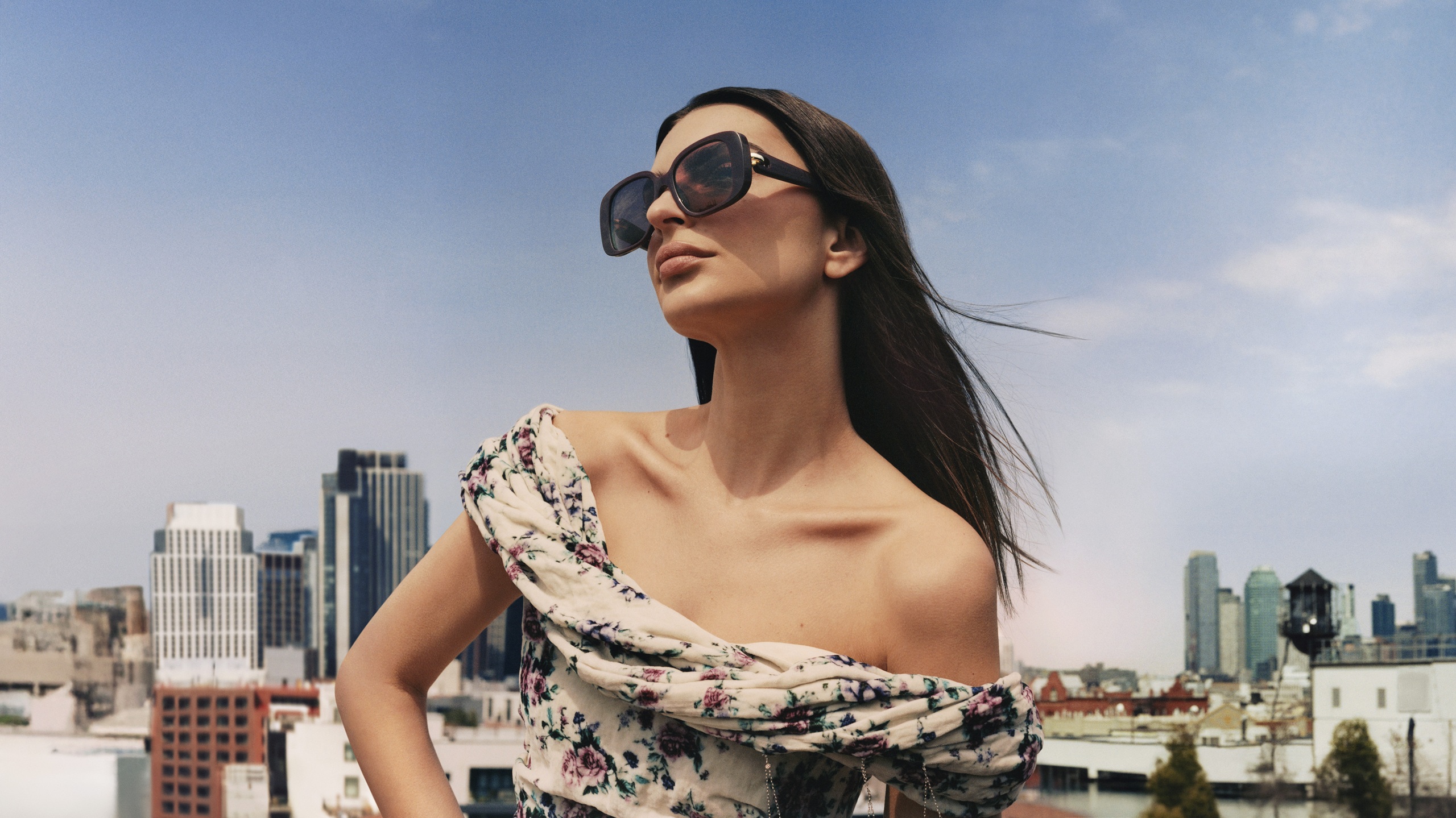 Emily Ratajkowski portant des lunettes de soleil Tory Burch "Pierced" noires avec des détails d'anneaux métalliques sur les branches, vêtue d'un haut asymétrique à motifs floraux, photographiée contre l'horizon de New York avec un ciel bleu et une architecture urbaine.