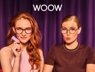 Two young women modeling WOOW eyewear against a purple curtain backdrop. The woman on the left has long red hair and wears tortoiseshell-patterned glasses while holding a purple card, dressed in a pink top. The woman on the right has blonde hair in a bun and wears round glasses with colorful accents, dressed in a brown sweater. The WOOW brand logo appears prominently at the top