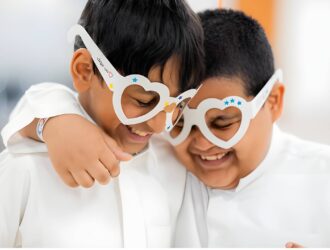 Deux jeunes garçons portant des chemises blanches s'embrassent en portant des montures de lunettes blanches en forme de cœur décorées d'étoiles bleues, souriant ensemble dans ce qui semble être un cadre de soins oculaires ou de dépistage visuel, représentant le thème 'Love Your Child's Eyes' du concours photo de la Journée mondiale de la vue.