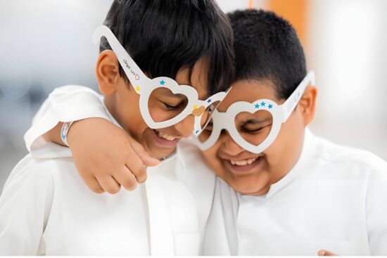 Deux jeunes garçons portant des chemises blanches s'embrassent en portant des montures de lunettes blanches en forme de cœur décorées d'étoiles bleues, souriant ensemble dans ce qui semble être un cadre de soins oculaires ou de dépistage visuel, représentant le thème 'Love Your Child's Eyes' du concours photo de la Journée mondiale de la vue.