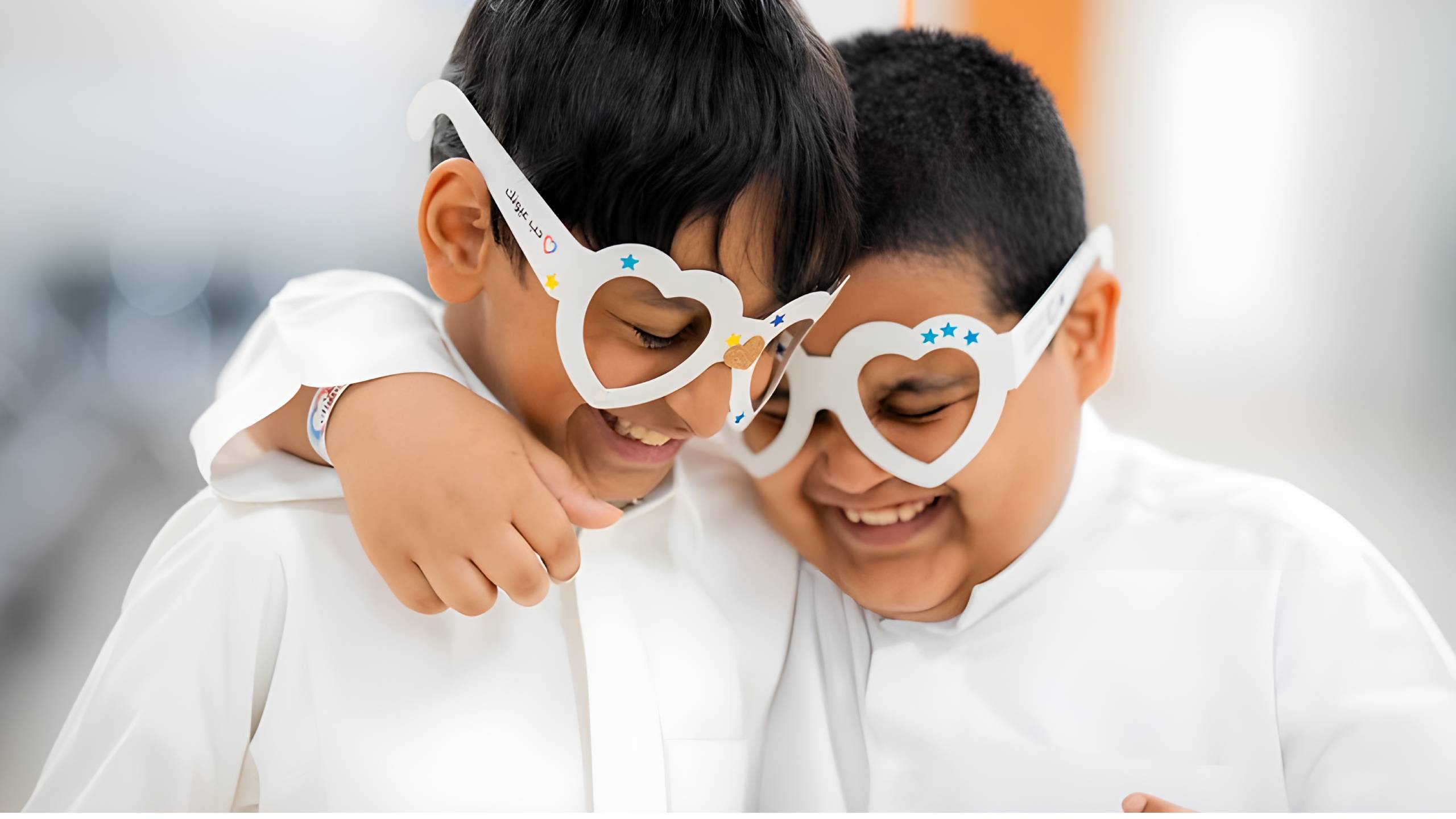 Two young boys wearing white shirts embrace while wearing heart-shaped white glasses frames decorated with blue stars, smiling together in what appears to be an eye care or vision screening setting, representing the 'Love Your Child's Eyes' theme of the World Sight Day Photo Competition.