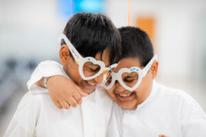 Deux jeunes garçons portant des chemises blanches s'embrassent en portant des montures de lunettes blanches en forme de cœur décorées d'étoiles bleues, souriant ensemble dans ce qui semble être un cadre de soins oculaires ou de dépistage visuel, représentant le thème 'Love Your Child's Eyes' du concours photo de la Journée mondiale de la vue.