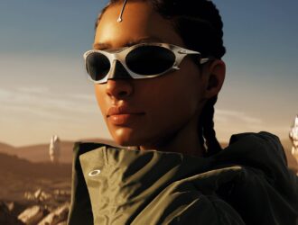 Close-up portrait of a woman wearing white wraparound sports sunglasses and olive green athletic wear, photographed in golden hour lighting against a desert landscape background with rocky formations visible in the distance.