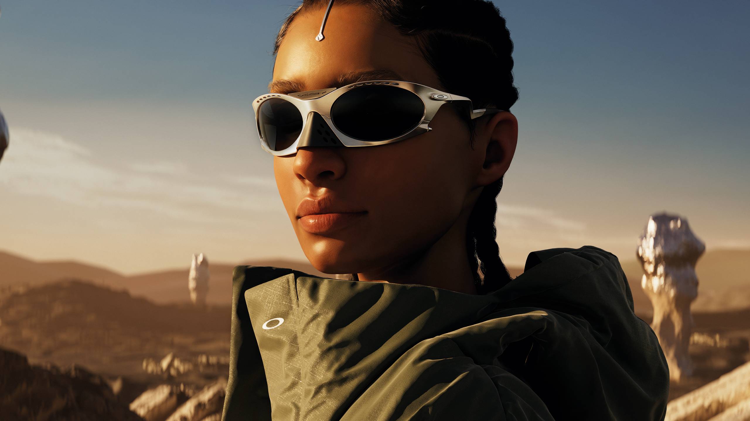 Portrait en gros plan d'une femme portant des lunettes de soleil de sport blanches enveloppantes et des vêtements de sport vert olive, photographiée à l'heure dorée contre un paysage désertique avec des formations rocheuses visibles au loin.