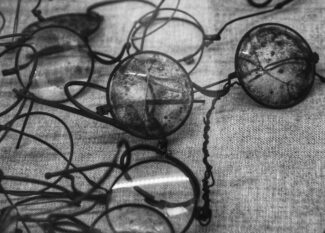 Photographie en noir et blanc montrant une collection de lunettes et de lunettes vintage détériorées dispersées sur une surface textile texturée. Les montures historiques présentent des signes visibles de vieillissement et de dégradation, avec certains verres paraissant troubles ou endommagés, illustrant les défis de conservation auxquels font face les matériaux de lunetterie en nitrate de cellulose et en acétate au fil du temps.