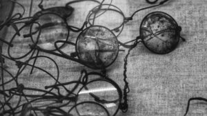 Black and white photograph showing a collection of deteriorated vintage eyeglasses and spectacles scattered on a textured fabric surface. The historic frames display visible signs of aging and degradation, with some lenses appearing cloudy or damaged, illustrating the preservation challenges faced by cellulose nitrate and acetate eyewear materials over time.