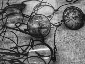 Black and white photograph showing a collection of deteriorated vintage eyeglasses and spectacles scattered on a textured fabric surface. The historic frames display visible signs of aging and degradation, with some lenses appearing cloudy or damaged, illustrating the preservation challenges faced by cellulose nitrate and acetate eyewear materials over time.