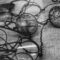 Black and white photograph showing a collection of deteriorated vintage eyeglasses and spectacles scattered on a textured fabric surface. The historic frames display visible signs of aging and degradation, with some lenses appearing cloudy or damaged, illustrating the preservation challenges faced by cellulose nitrate and acetate eyewear materials over time.
