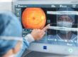 English: "An eye care professional in a white coat pointing at a retinal scan displayed on a computer monitor, showing an orange-colored fundus image of the back of an eye with targeting crosshairs, while a patient wearing a face mask is visible in the background.